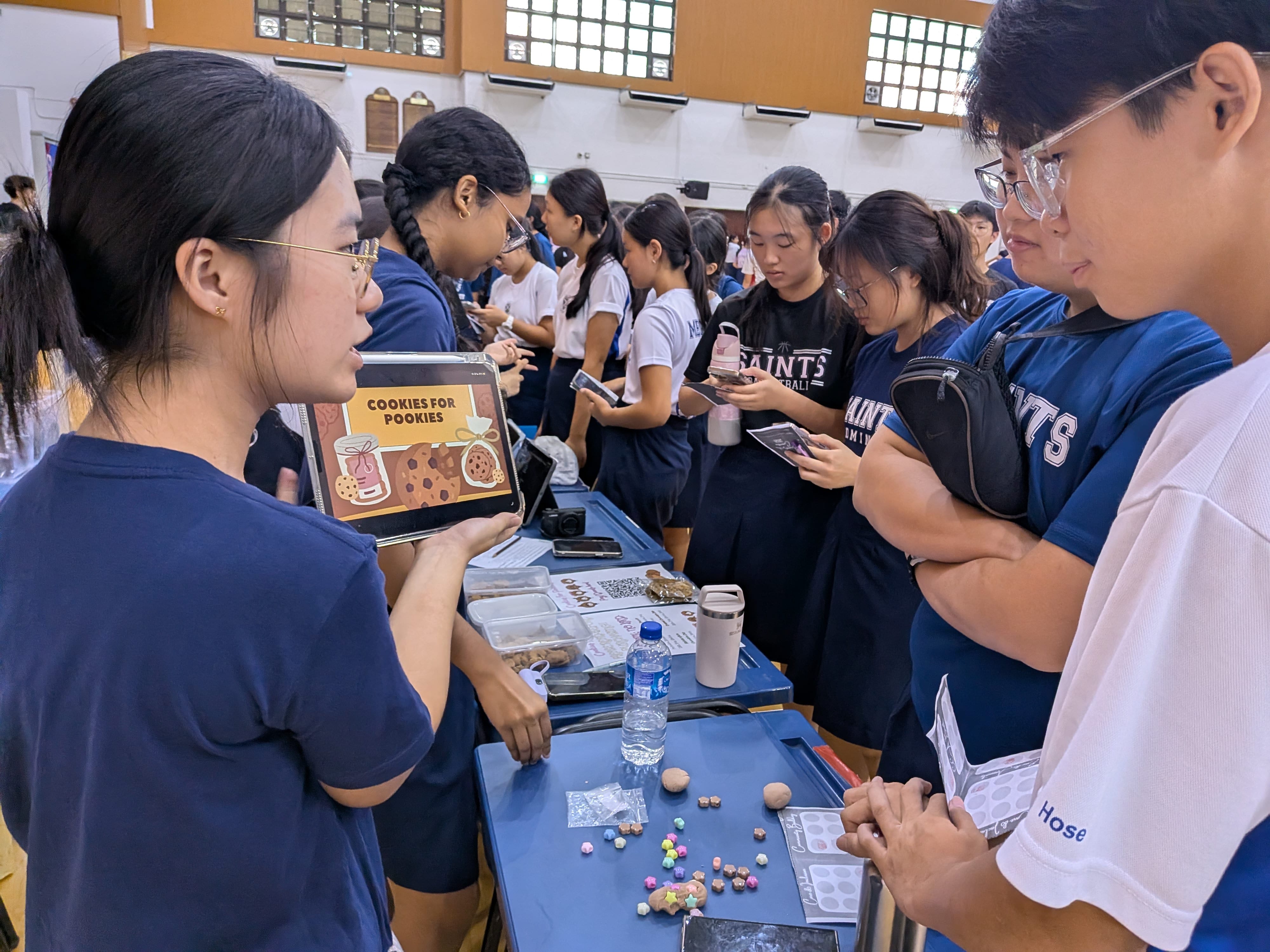 A student presenting a digital slide titled "Cookies for Pookies" on a tablet to peers gathered around an interactive exhibition booth.
